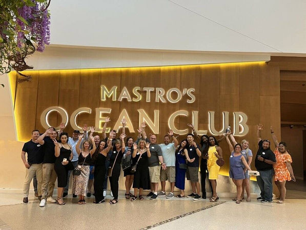 Group photo of about 20 adults smiling and waving in front of a large illuminated ocean-themed restaurant sign on a warm wood wall, casual evening attire at an indoor entrance.