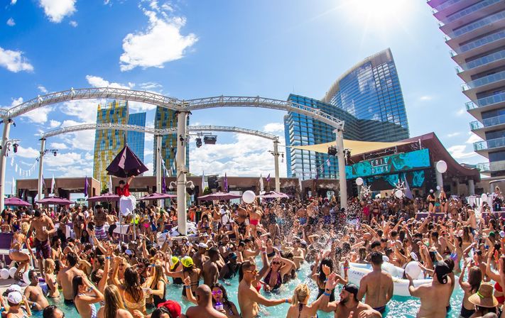 Crowded daytime Las Vegas pool party at an outdoor dayclub with people dancing and splashing in the pool, DJs and stage, purple umbrellas, and high-rise glass hotels under a bright blue sky