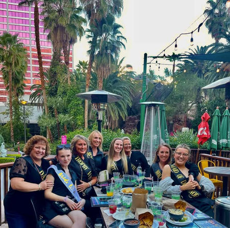 Eight women wearing sashes, including a bride‑to‑be, celebrating a bachelorette party on a palm‑lined outdoor restaurant patio with string lights, patio heaters, drinks and snacks.