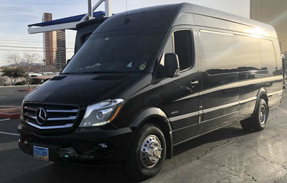 Sleek black Mercedes Sprinter high-roof van parked in a sunlit commercial parking lot with a city tower and low hills in the background.
