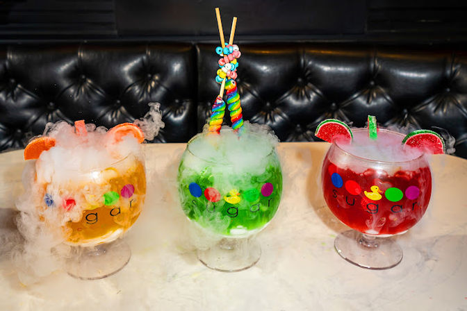 Three colorful dry-ice cocktails on a marble table in a black leather lounge, garnished with citrus slices, gummy candies and rainbow lollipops