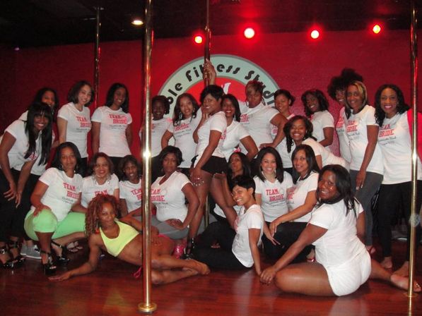Bachelorette party group wearing 'Team Bride' T-shirts posing around dance poles in a red-lit pole fitness studio, women smiling and striking playful dance poses.
