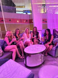 Bachelorette party of five women in cocktail dresses wearing sashes, toasting martinis on curved sofas in a neon-pink modern hotel lounge with hanging bead curtains and a round marble table.