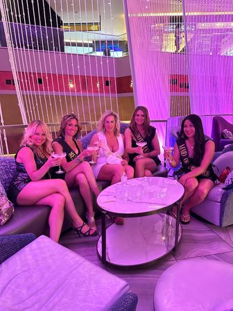 Bachelorette party of five women in cocktail dresses wearing sashes, toasting martinis on curved sofas in a neon-pink modern hotel lounge with hanging bead curtains and a round marble table.