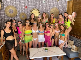 Large group of women in colorful swimwear and cover-ups smiling and posing in a sunny poolside cabana with woven circular wall decor, round mirror, wooden accents and a marble-top table with a small succulent — tropical beach club vibe.