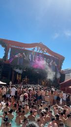 Crowded Las Vegas daytime pool party with people in swimsuits packed in the pool and on the deck, DJ performing on a large stage with LED screen, dancers and smoke under a clear blue sky.