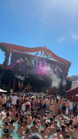 Crowded Las Vegas daytime pool party with people in swimsuits packed in the pool and on the deck, DJ performing on a large stage with LED screen, dancers and smoke under a clear blue sky.