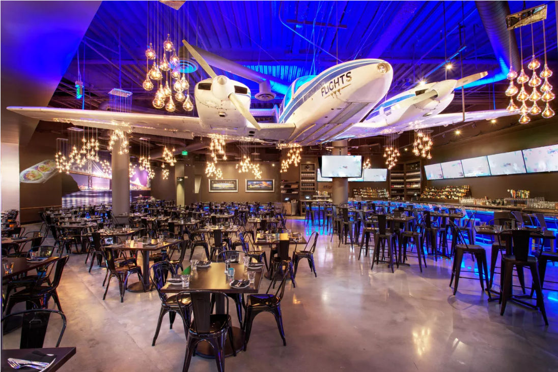 Aviation-themed indoor restaurant and bar with a twin-engine airplane suspended from the ceiling, Edison bulb cluster lighting, blue accent lights, rows of metal tables and chairs and a polished concrete floor.