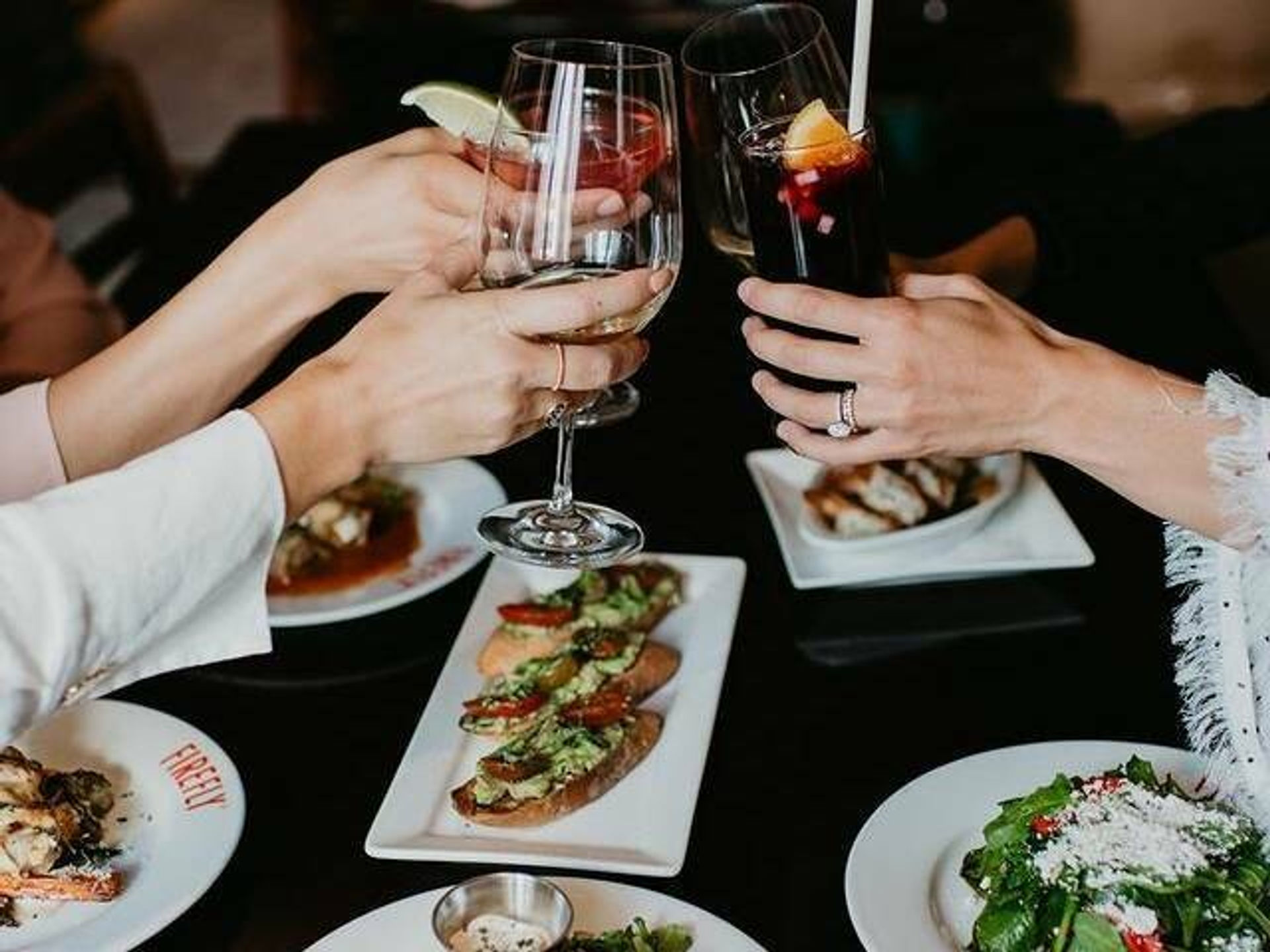 Three hands clinking wine glasses and a cocktail over a bistro table of avocado bruschetta, mixed green salad and small plates — casual restaurant cheers