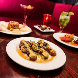 Grilled meat skewers drizzled with creamy green sauce on a white plate at a dimly lit restaurant table, with cocktails and small plates blurred in the background.