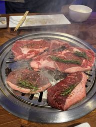 Sizzling steaks on a round tabletop Korean BBQ grill — five seasoned raw beef cuts topped with rosemary and pepper flakes, steam rising; chopsticks and rice bowl on wooden table in background.