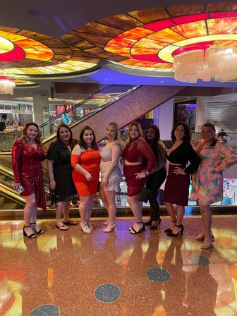 Eight women in evening dresses posing on a shiny hotel/casino lobby floor under a colorful stained-glass ceiling and chandelier, escalators visible in the background.