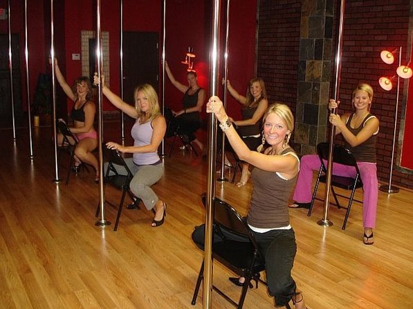 Group of smiling women in a pole fitness class in a bright dance studio with wooden floors, metal poles, chairs and red walls.