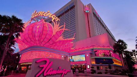 Pink neon-lit casino hotel façade at dusk with a giant lotus-shaped marquee, glowing signage, and palm trees along a lively Las Vegas-style strip.