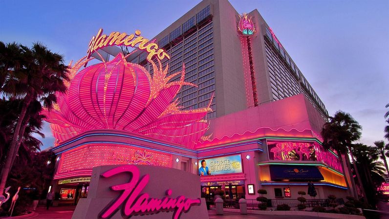 Pink neon-lit casino hotel façade at dusk with a giant lotus-shaped marquee, glowing signage, and palm trees along a lively Las Vegas-style strip.