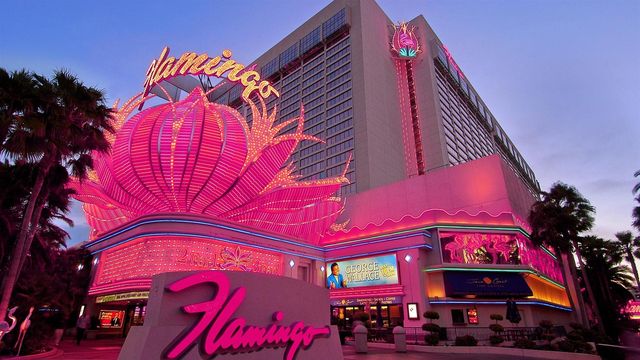Pink neon-lit casino hotel façade at dusk with a giant lotus-shaped marquee, glowing signage, and palm trees along a lively Las Vegas-style strip.