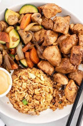 Savory teriyaki chicken bowl with cubed glazed chicken, zucchini-carrot-mushroom stir-fry, seasoned fried rice, dipping sauce and chopsticks