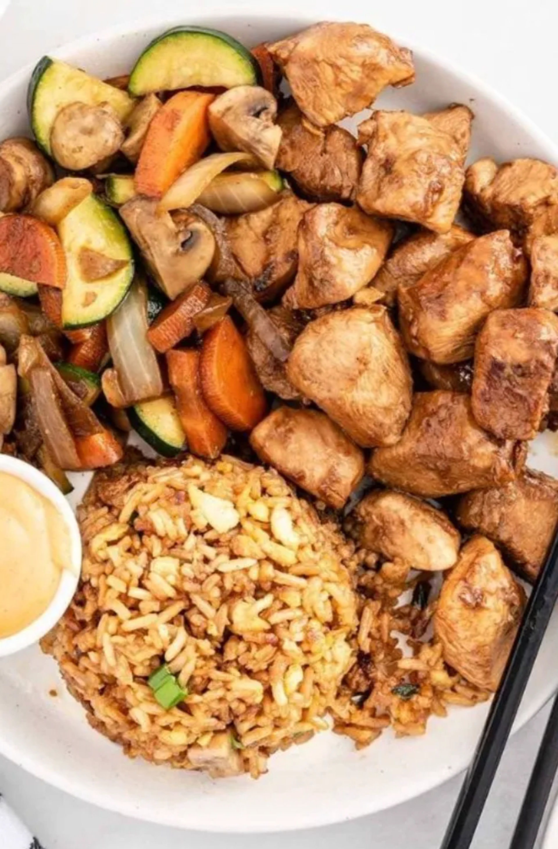 Savory teriyaki chicken bowl with cubed glazed chicken, zucchini-carrot-mushroom stir-fry, seasoned fried rice, dipping sauce and chopsticks
