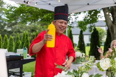 Smiling chef in a red jacket and tall black hat holds a yellow squeeze bottle under a white canopy at a backyard garden catering event with floral table centerpieces and evergreen hedges in the background.