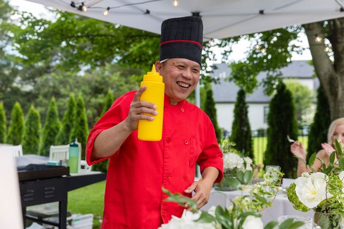 Smiling chef in a red jacket and tall black hat holds a yellow squeeze bottle under a white canopy at a backyard garden catering event with floral table centerpieces and evergreen hedges in the background.