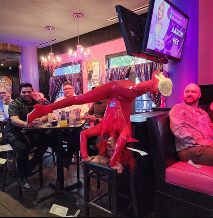 Drag performer in shiny red latex and platform boots doing an upside-down split on a bar stool in a pink-lit cabaret-style bar as patrons at a nearby table watch and toss dollar bills.