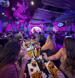 Purple neon-lit restaurant bar with a stage and disco balls, crowded long table of friends enjoying burgers, fries and drinks during live entertainment