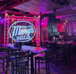 Neon-lit Dallas bar interior with small stage framed by metal truss and circular logo projection, high-top tables and chairs, and purple-pink mood lighting for live-music nightlife.