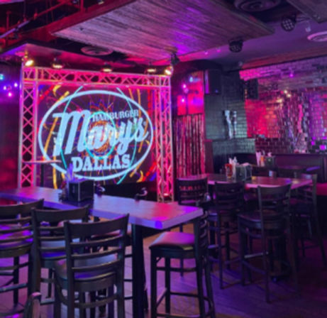 Neon-lit Dallas bar interior with small stage framed by metal truss and circular logo projection, high-top tables and chairs, and purple-pink mood lighting for live-music nightlife.