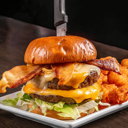 Stacked double bacon cheeseburger on a glossy brioche bun with two melted cheese layers, crispy bacon, shredded lettuce and a knife through the top, served with seasoned tater tots on a white rectangular plate.