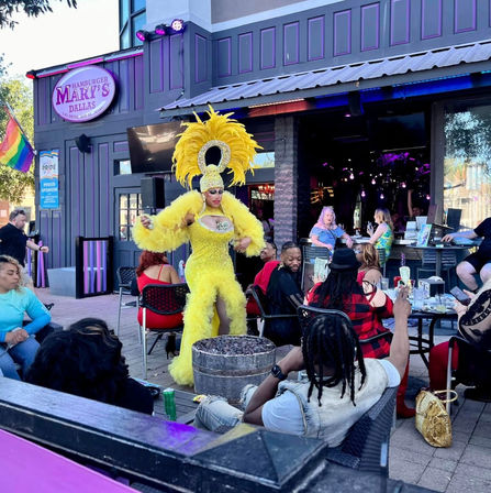 Drag performer in a sparkling yellow feathered costume and towering headdress entertains a lively outdoor patio crowd beneath a Pride flag.