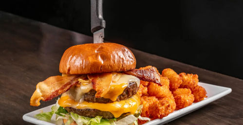 Towering bacon double cheeseburger with melted cheddar on a brioche bun, knife stuck through the top, served with crispy tater tots on a white plate on a wooden table