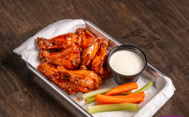 Crispy buffalo chicken wings glazed in bright hot sauce on a parchment-lined metal tray with ranch dipping sauce and celery and carrot sticks on a rustic wooden table
