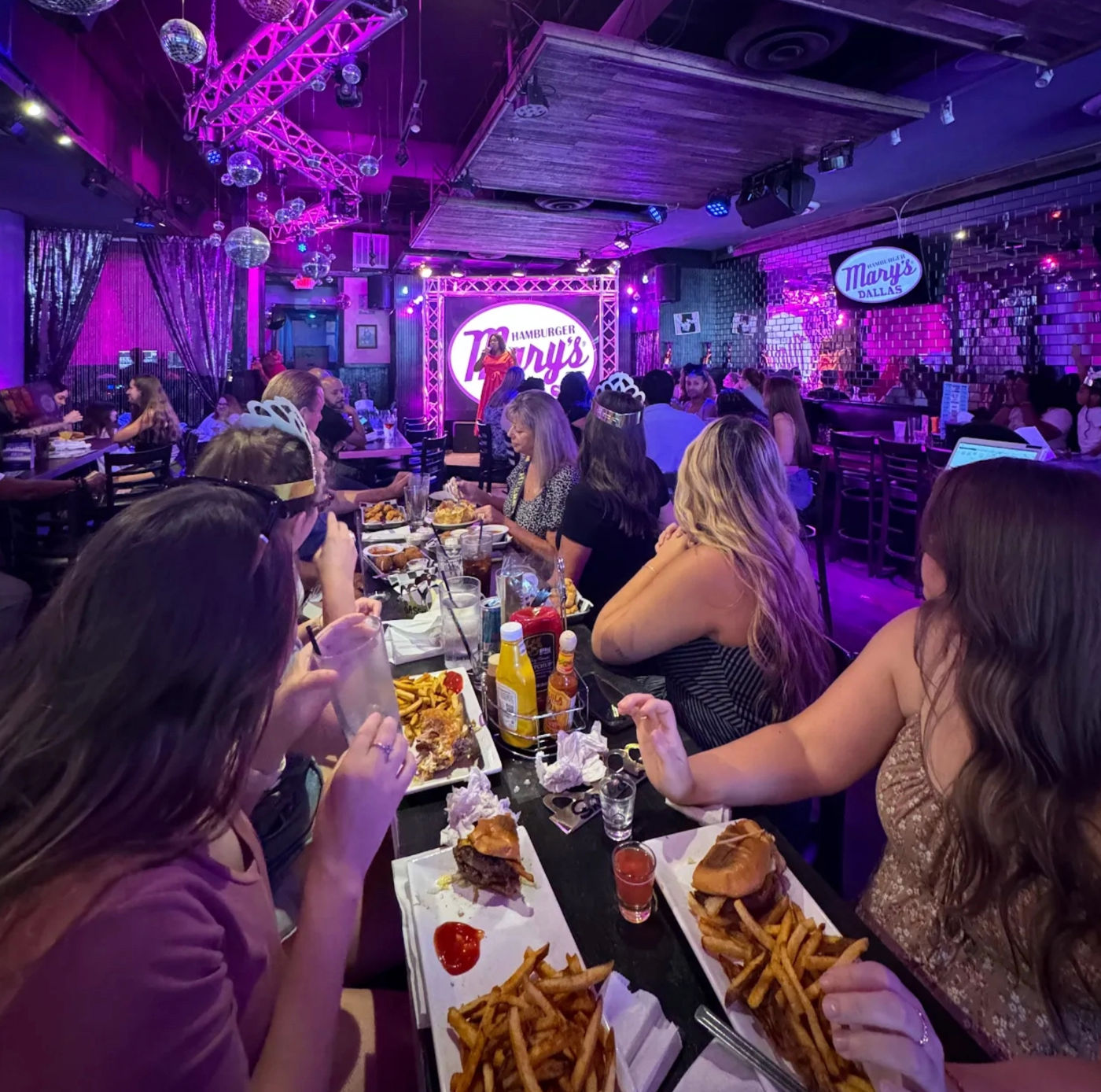 Neon-lit burger restaurant and bar with a live performer on stage, disco balls overhead, and a lively table of friends enjoying burgers and fries.