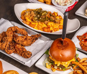 Overhead shot of American comfort-food spread: cheeseburger with melted cheddar and a knife through a brioche bun, crispy fried chicken tenders, loaded cheesy tater tots topped with jalapeños, golden fries and coleslaw in a casual bar-and-grill style meal.