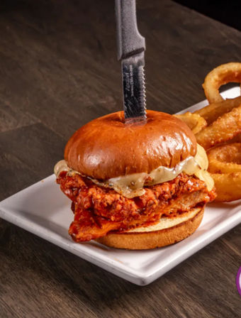 Crispy spicy fried chicken sandwich with melted cheese on a glossy brioche bun skewered by a steak knife, served with golden onion rings on a white plate over a dark wood table