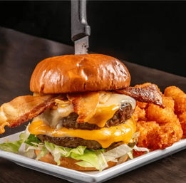 Mouthwatering stacked double bacon cheeseburger with melted cheddar and Swiss, shredded lettuce on a glossy brioche bun with a knife through the top, served with crispy tater tots on a white plate.