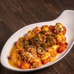 Crispy loaded tater tots smothered in nacho cheese and seasoned ground beef, topped with pickled jalapeños and shredded cheddar in a white oval dish on a wooden table, Tex‑Mex appetizer