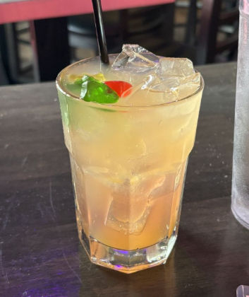 Iced orange-yellow cocktail in a tall glass with ice, black straw and red and green gummy candy garnish on a dark wooden bar table in an indoor bar