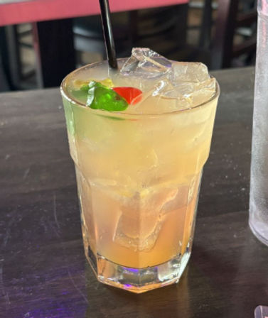 Iced orange-yellow cocktail in a tall glass with ice, black straw and red and green gummy candy garnish on a dark wooden bar table in an indoor bar