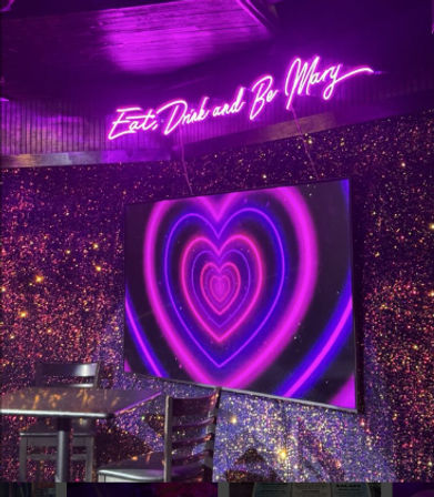 Neon-lit lounge interior with purple script sign reading 'Eat, Drink and Be Merry' above a glitter-speckled wall and a large screen showing concentric pink and purple neon hearts, with a metal table and chairs in the foreground.
