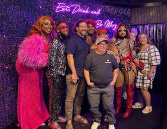 Glittery nightclub group shot of drag performers in sequins, feathers and thigh-high boots posing with smiling guests on a sparkly stage beneath a neon sign reading Eat Drink and Be Merry