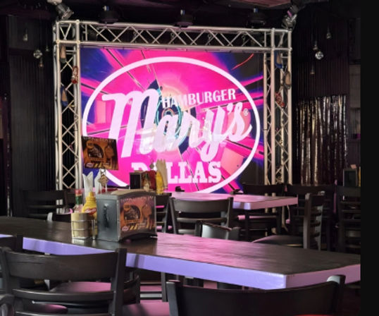 Empty restaurant/bar interior with wooden tables and condiment caddies, colorful pink circular stage sign framed by truss lighting — lively Dallas vibe.