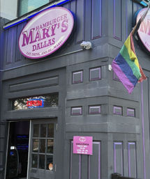 Exterior of a gray-and-purple corner burger bar in Dallas with a pink round sign, neon OPEN window and a waving rainbow pride flag.