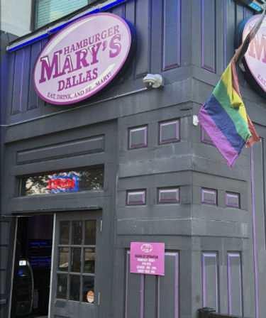 Exterior of a gray-and-purple corner burger bar in Dallas with a pink round sign, neon OPEN window and a waving rainbow pride flag.