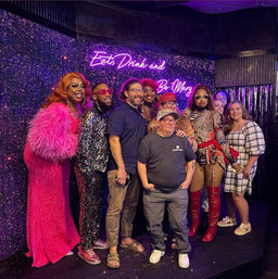 Lively drag show group photo on a glittery stage with a purple neon sign reading “Eat Drink and Be Mary,” featuring performers in sequins, feathers, bold makeup and colorful costumes posing with smiling audience members.
