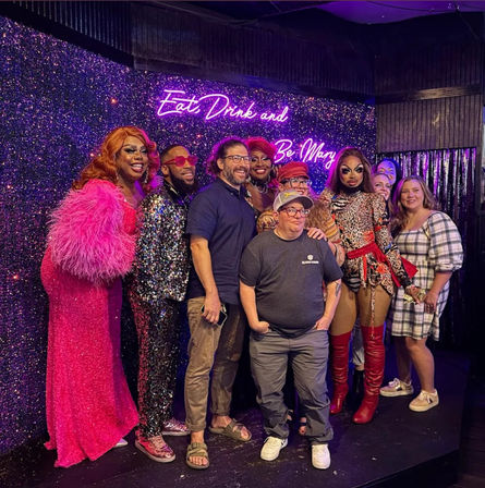 Lively drag show group photo on a glittery stage with a purple neon sign reading “Eat Drink and Be Mary,” featuring performers in sequins, feathers, bold makeup and colorful costumes posing with smiling audience members.