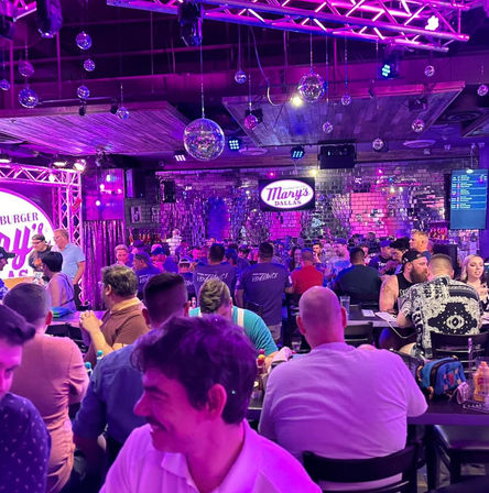 Lively neon-lit bar scene in Dallas, packed with people at long tables under hanging disco balls and purple-pink stage lighting against a mirrored wall.