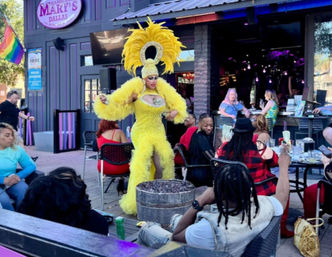 Drag performer in a towering yellow feathered headdress and sparkling gown entertaining a lively outdoor patio crowd at a Dallas bar, Pride flag visible and guests seated at tables.