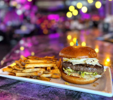 Juicy cheeseburger on a brioche bun with lettuce and melted cheese, served with golden fries on a white plate at a colorful indoor bar with purple bokeh lights.
