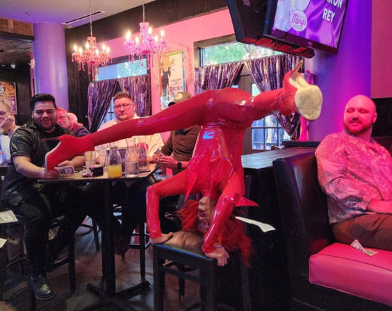 Contortion performer in shiny red latex and matching heels doing a handstand across a bar stool in a pink‑lit cabaret bar while nearby patrons at a table toss cash.